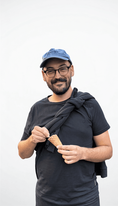 Mann mit Brille und blauer Kappe, hält eine Eistüte in der Hand und lächelt in die Kamera.
