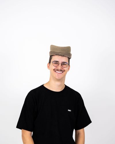 Person mit schwarzem T-Shirt und beige Hut, lächelnd mit Händen in den Hosentaschen, vor weißem Hintergrund.
