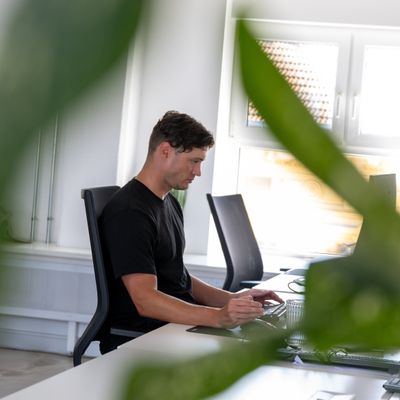 Mann in schwarzem T-Shirt arbeitet konzentriert an einem Laptop in einem modernen Büro, umgeben von grünen Pflanzen.