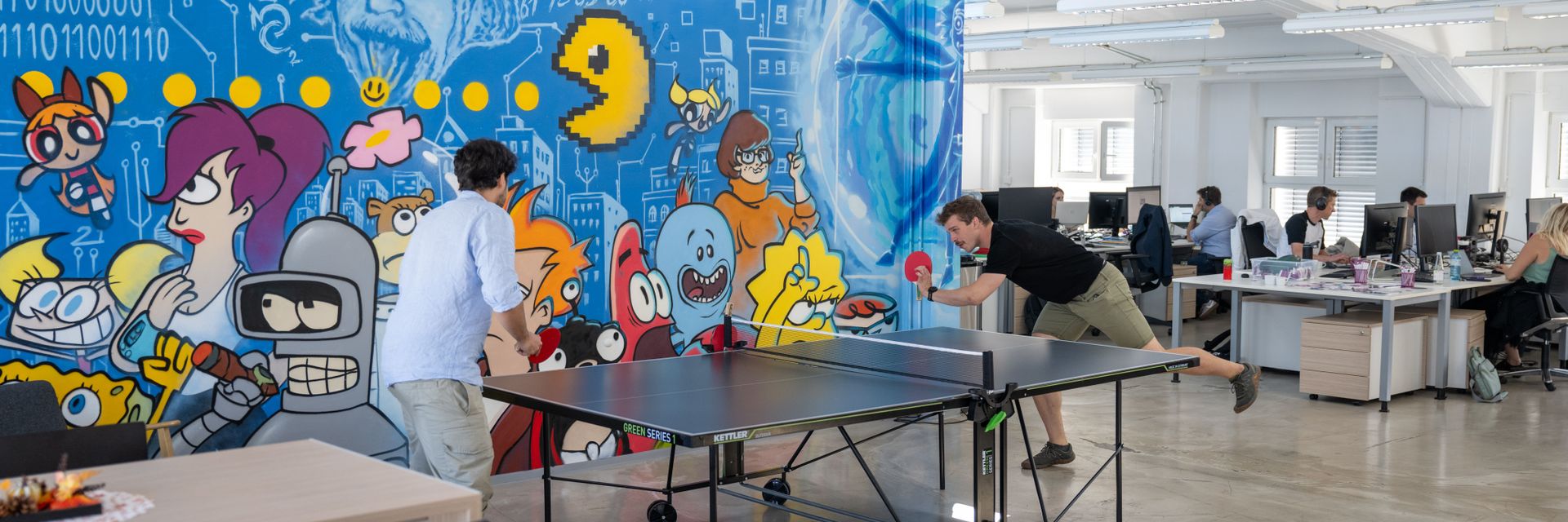 Zwei Personen spielen Tischtennis in einem Büro mit buntem Wandgemälde von Cartoon-Figuren. Im Hintergrund arbeiten Mitarbeiter an Schreibtischen.