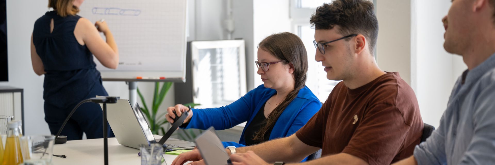 Kollegen in einem Meetingraum, die an Laptops arbeiten, während eine Frau am Whiteboard präsentiert.