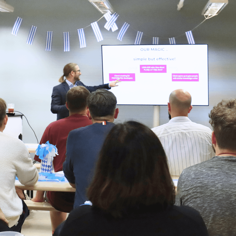 Person hält eine Präsentation vor einer Gruppe von Menschen, die auf Bänken sitzen, während auf einem Bildschirm eine Folie mit Text angezeigt wird.